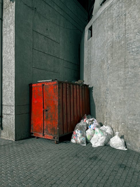 A red metal waste container with visible rust and peeling paint is situated against a dark grey stone wall on the left and a plain concrete wall on the right. The container is rectangular with vertical ridges along its sides and is partially closed, with a small latch on the front. Surrounding the base of the container are several clear plastic garbage bags filled with mixed household waste, some with visible crumples and folds. The scene is outdoors, on a textured grey paving surface composed of small, interlocking pattern bricks. The background includes a dark building façade with smooth paneling, and a small, square window is visible on the upper part of the concrete wall. The overall atmosphere is neutral and functional, reflecting a typical outdoor rubbish disposal area where private waste collection occurs, aligning with independent rubbish removal services such as those offered by Junk Removal Services.