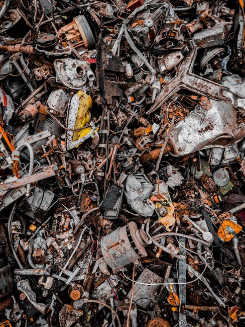 A large pile of assorted automotive scrap metal and parts, including engine components, gears, and small mechanical fragments, all rusted and weathered with various shades of brown, orange, and grey. The items are jumbled together on a concrete or asphalt surface, with some pieces visibly corroded and covered in dirt or grease. The background shows no clear environment but suggests an outdoor or industrial setting, with the scrap material occupying most of the frame. The scene reflects a disorderly collection of discarded vehicle parts, illustrating the type of waste that might be managed through independent collection or off-site clearance services, such as those offered by Junk Removal Services, to facilitate proper disposal or recycling of automotive materials.